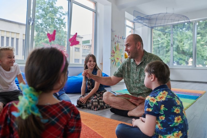 Male teacher teaching a group of young children, all sitting on the floor in a brightly lit classroom.