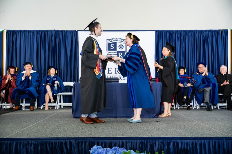 Dever receiving his diploma from President Armstrong in full regalia, with blue drapery and a Salve sign in the background.
