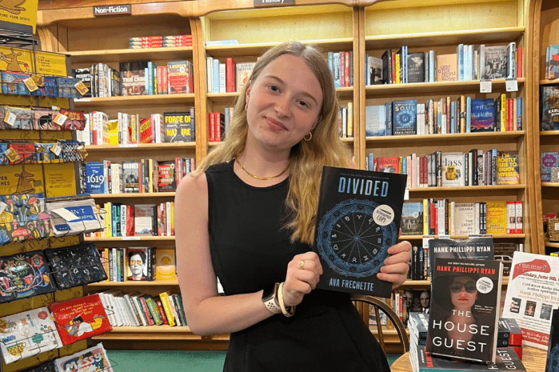 Ava wearing a black dress in a bookstore holding a copy of her first book "Divided"