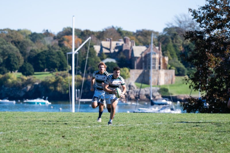 two men's rugby players on the field running after ball with ocean in background.