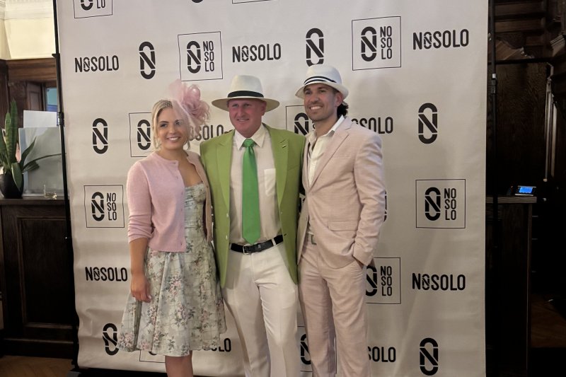 group photo of three people in pink and green outfits in front of a NOSOLO backdrop, for a Kentucky Derby fundraising event.