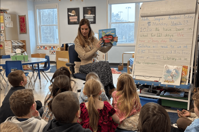 Teacher reads a picture book to students seated on a classroom rug.