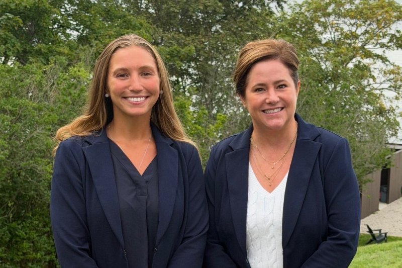 Two women stand together outdoors with Newport Harbor in the background.