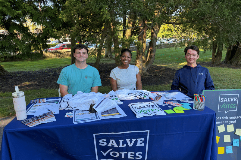 Three SalveVotes student members sit behind a voter engagement table outdoors on campus.