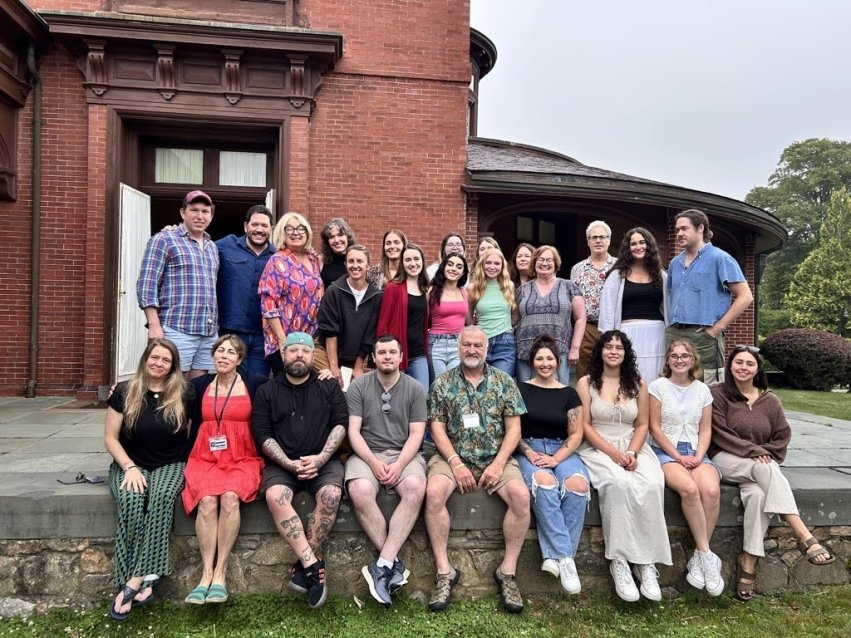 A group photo with the back row standing and front row sitting on a ledge, smiling in front of a red building.