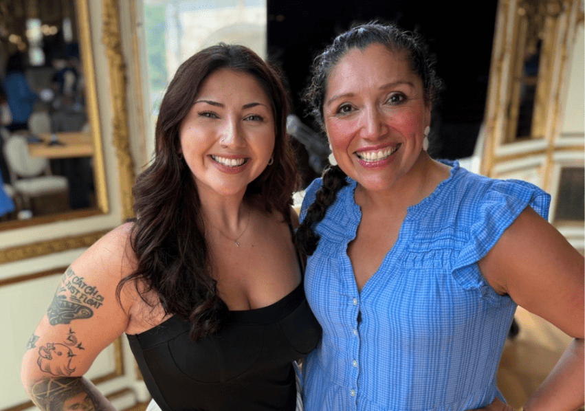 Acevedo (left) in a black top and Jennifer De Leon in a blue dress together and smiling at the camera.