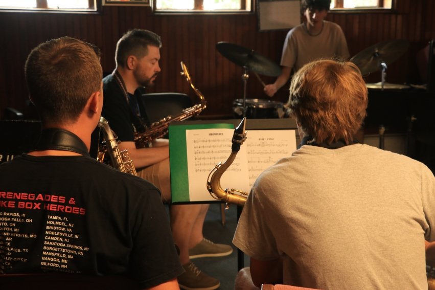 Students with their backs to the camera playing the saxophone while reading music placed on their music stand.