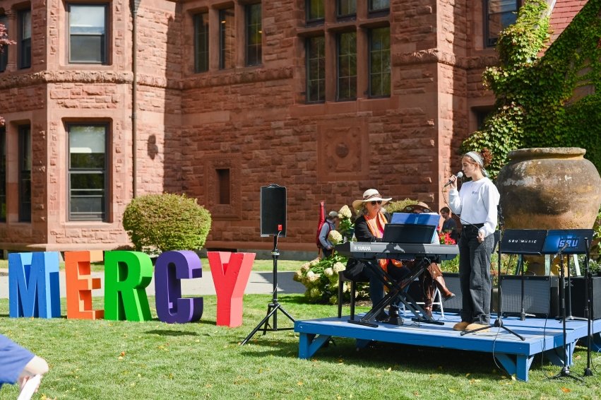Student singing on stage outside next to "MERCY" sign.