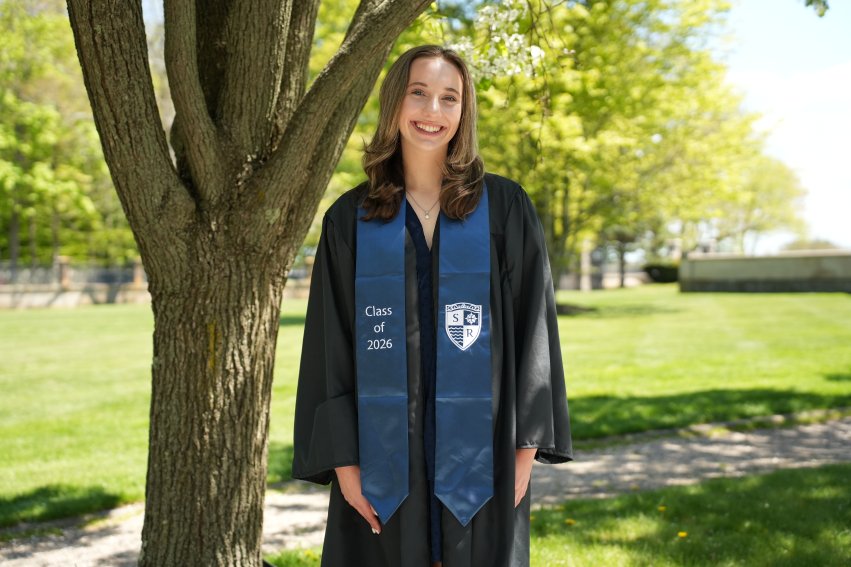 augustine in graduation regalia smiling in front of tree
