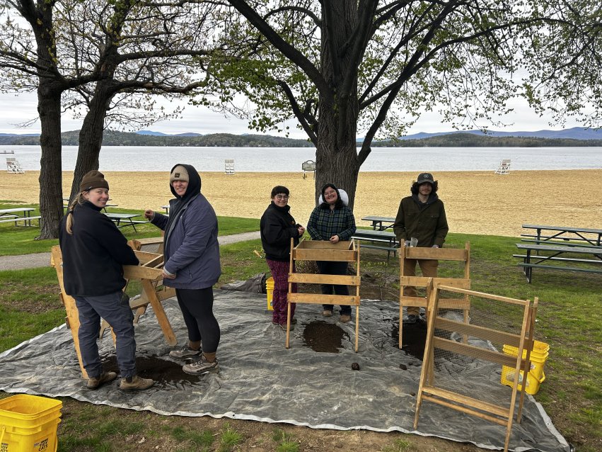 Students use wooden soil screens to sift archaeological material beside a lakeshore beach during archaeological field school.