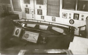 Black-and-white postcard of a wooden lifeboat displayed indoors in a museum gallery.