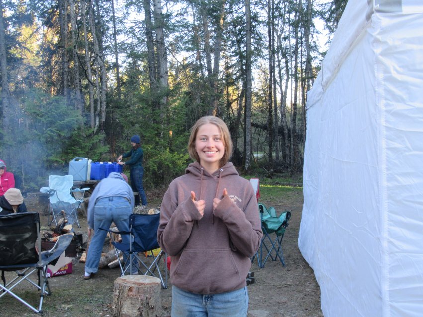 Female student at an archaeological field camp in a forested area, smiling and giving two thumbs up while classmates prepare camp in the background.