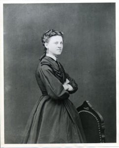 Black-and-white portrait of a young Ida Lewis standing in profile with her arms crossed beside an upholstered chair, wearing a long-sleeved, high-necked dress.