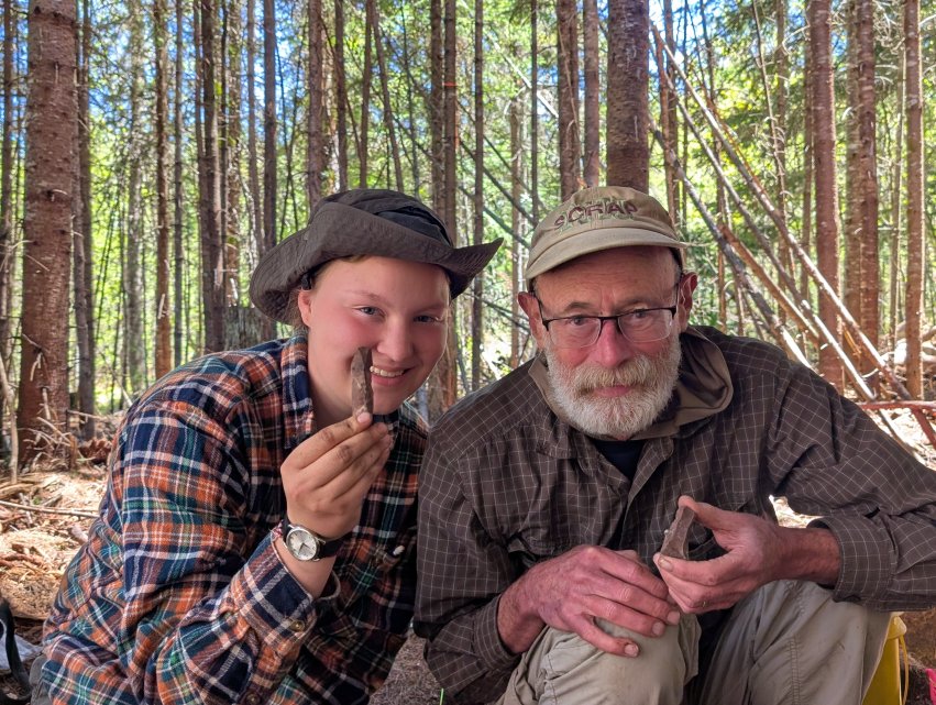Cultural and historic preservation student and instructor kneel in a forest, each holding a stone tool recovered during fieldwork in Maine.