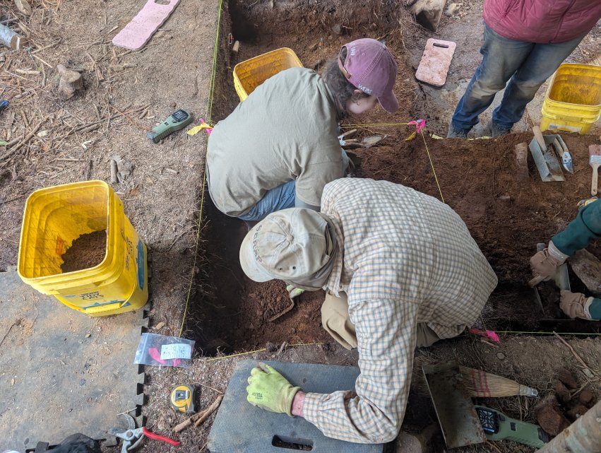 Cultural and historic preservation student and instructor carefully excavate an archaeological unit using hand tools during fieldwork in Maine.