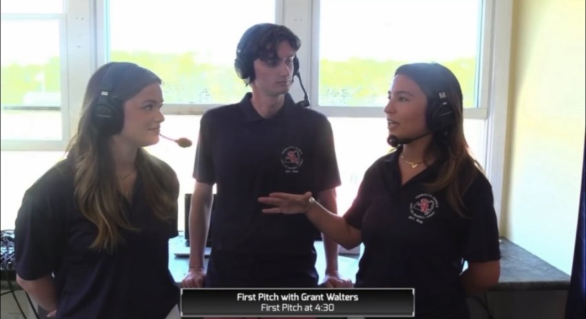 Three people wearing headsets broadcast a baseball game from the press box.