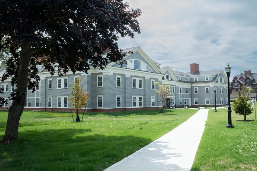 A large gray-shingled building with white trim, surrounded by lawns, trees and a paved walkway.