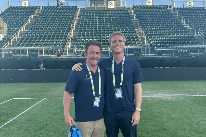 Two staff members pose on a grass court at the International Tennis Hall of Fame with the stadium seating behind them.