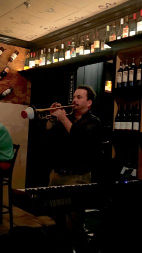 Luke Bayne performs the trumpet beside a keyboard in a dimly lit restaurant, with shelves of wine bottles lining the walls behind him.