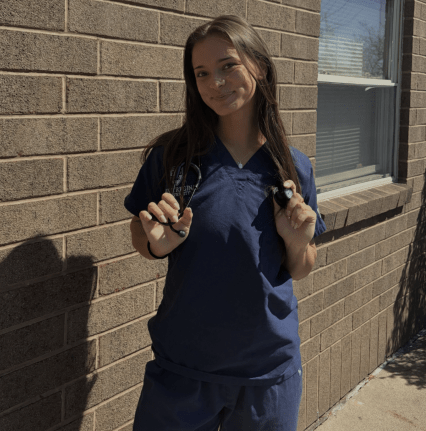Belair in her navy blue nursing scrubs smiling with a stethoscope around her neck.